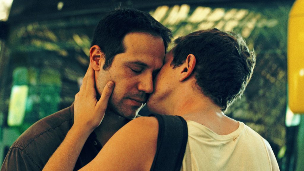 A still from PRIVATE DESERT: A man holds another man with tenderness and kisses his cheek.